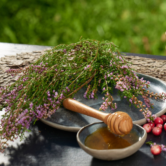 Flowering heather sprigs on a grey plate, a honey dipper resting in a small bowl of honey, red berries nearby