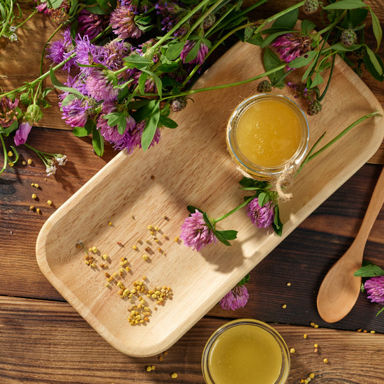 Red clover flowers, bee pollen, a wooden spoon and two small jars of honey on a wooden table